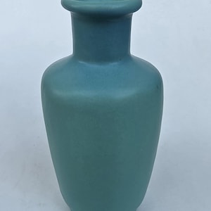 May include: A matte teal ceramic vase with a smooth, rounded body and a flared rim. The vessel features a consistent solid color finish across its entire surface, showcasing a simple, functional design with a narrow neck and a stable base.