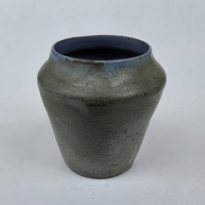 May include: A handcrafted ceramic vase with a wide, slightly flared body and a dark blue interior. The exterior has a mottled gray-green glaze with a light blue rim. The vase has a rustic, organic aesthetic.