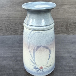 May include: A ceramic vase with a flared rim and a cylindrical body. The vase features a light blue and cream speckled glaze with a painted design of blue curved lines, a small red dot, and a blue flower motif near the base of the vessel.