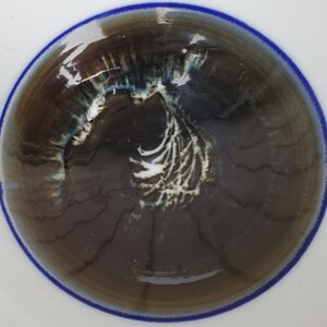 May include: A close-up view of a ceramic bowl interior. The bowl's interior features a dark brown and gray swirling pattern with white accents. The rim is white with a thin blue border. The bowl's design is abstract and artistic.