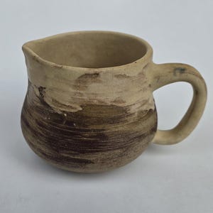 May include: A small ceramic creamer pitcher featuring a rounded body with a wide handle and a pouring spout. The exterior displays a two-tone design with a light beige upper section and a dark brown, textured lower section with visible brush strokes.