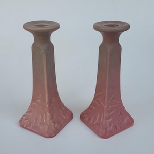 May include: Two matching, dusty rose-colored candle holders. Each has a square base with leaf-like detailing, a tall, rectangular body, and a circular top. The candle holders are made of ceramic or a similar material and have a matte finish.