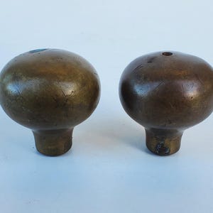 May include: Two antique brass door knobs with a round shape and a stem. The knobs have a dark, aged patina, showing signs of wear. The knobs are likely vintage or repurposed hardware.