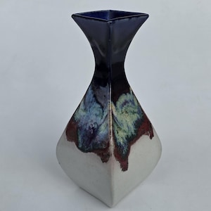 May include: A ceramic vase with a square top and a flared neck. The top is dark blue, transitioning to a white base. The middle section features a blend of blue, green, and maroon colors. The vase has a unique, geometric shape.