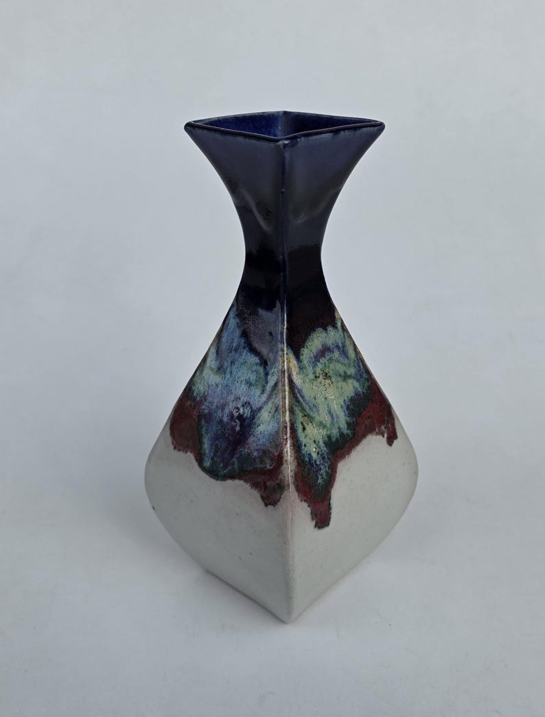May include: A ceramic vase with a square top and a flared neck. The top is dark blue, transitioning to a white base. The middle section features a blend of blue, green, and maroon colors. The vase has a unique, geometric shape.