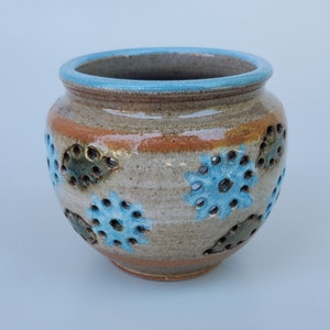 May include: A small, round, ceramic pot with a light brown glaze and blue and green floral designs. The pot has holes around the sides.