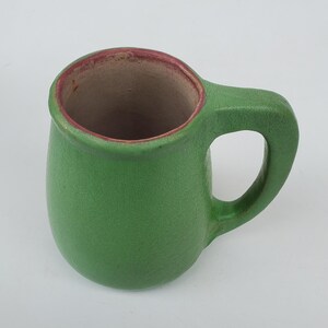 May include: A green ceramic mug with a rounded body and a handle. The interior is a light brown color, and the rim has a reddish-brown accent. The mug is designed for beverages and is suitable for home use.