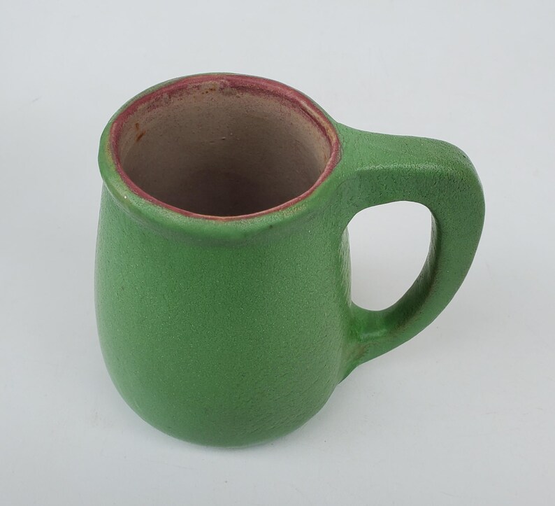 May include: A green ceramic mug with a rounded body and a handle. The interior is a light brown color, and the rim has a reddish-brown accent. The mug is designed for beverages and is suitable for home use.