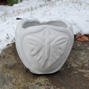 May include: White ceramic planter shaped like a heart with a butterfly design. The planter is perfect for small plants or succulents.