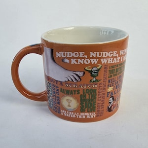 May include: A brown ceramic mug featuring various colorful illustrations and printed text including phrases like Nudge Nudge Wink Wink, Always Look on the Bright Side of Life, and Nobody Expects the Spanish Inquisition, with a white interior.