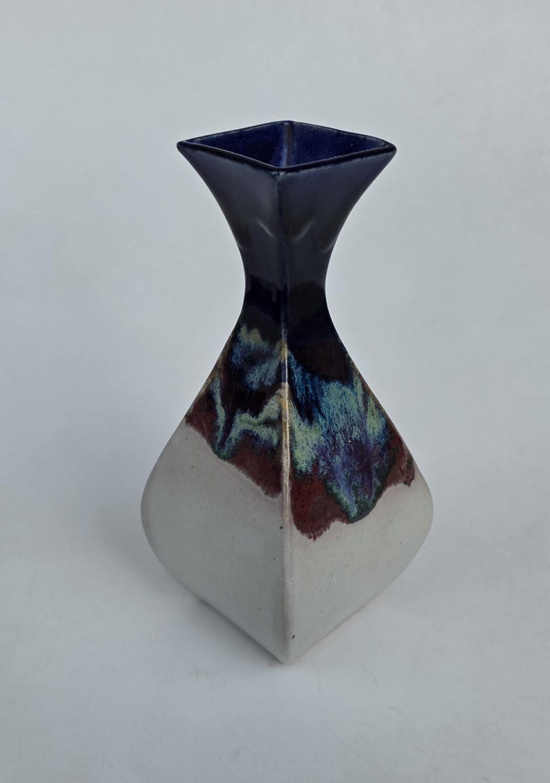 May include: A ceramic vase with a unique shape and color scheme. The top is a deep navy blue, transitioning to a blend of blue, green, and maroon. The base is a soft white, creating a striking contrast. This decorative vase is ideal for home decor.