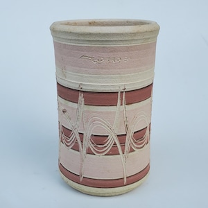 May include: A cylindrical ceramic vase with a light pink and maroon striped design. The vase has a textured, off-white rim and base. White, wavy lines are etched into the pink stripes. The word "CORRE" is etched into the pink band near the top.