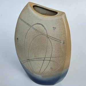 May include: A ceramic vase with a flattened oval shape, featuring a speckled beige upper section and a dark blue base. The surface is decorated with etched geometric lines and small circular patterns, with a wide, narrow opening at the top.