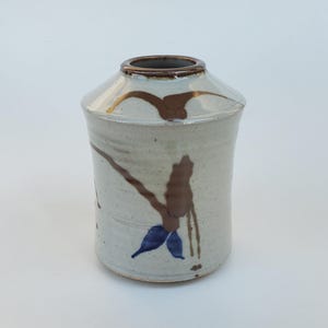 May include: A ceramic vase with a light beige base color, featuring brown and blue abstract painted designs. The vase has a cylindrical body with a slightly flared top and a dark brown rim. The overall design is rustic and artistic.