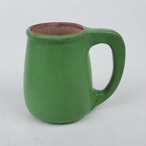 May include: A small, green ceramic mug with a rounded body and a large handle. The interior rim is a reddish-brown color. The mug has a textured surface and is likely handmade. This mug is suitable for coffee, tea, or other beverages.