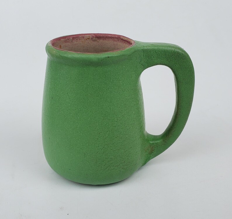 May include: A small, green ceramic mug with a rounded body and a large handle. The interior rim is a reddish-brown color. The mug has a textured surface and is likely handmade. This mug is suitable for coffee, tea, or other beverages.