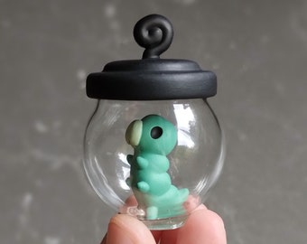 May include: A small glass jar with a black lid and a green and white caterpillar inside. The jar is being held in a hand.