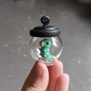 Op de afbeelding: Een klein glazen potje met een zwart deksel en een groene en witte rups erin. Het potje wordt in een hand gehouden.