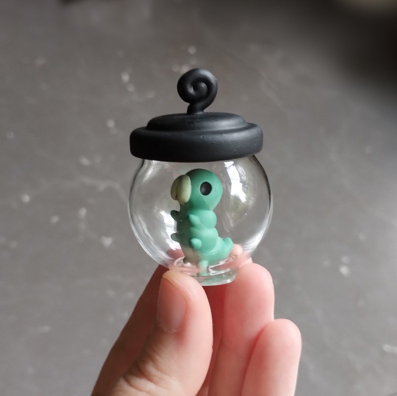 Op de afbeelding: Een klein glazen potje met een zwart deksel en een groene en witte rups erin. Het potje wordt in een hand gehouden.