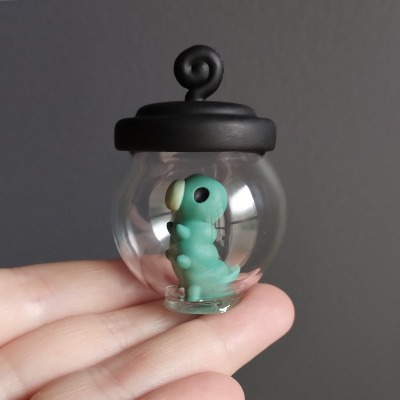 Op de afbeelding: Een klein glazen potje met een zwart deksel en een groene rupsfiguur erin. Het potje heeft een kleine lus bovenop om op te hangen.