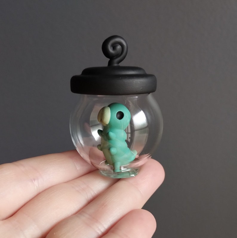 Op de afbeelding: Een kleine, heldere glazen pot met een zwart deksel en een zwarte swirl aan de bovenkant. In de pot zit een teal-kleurig, handgemaakt, kleifiguur van een rups.