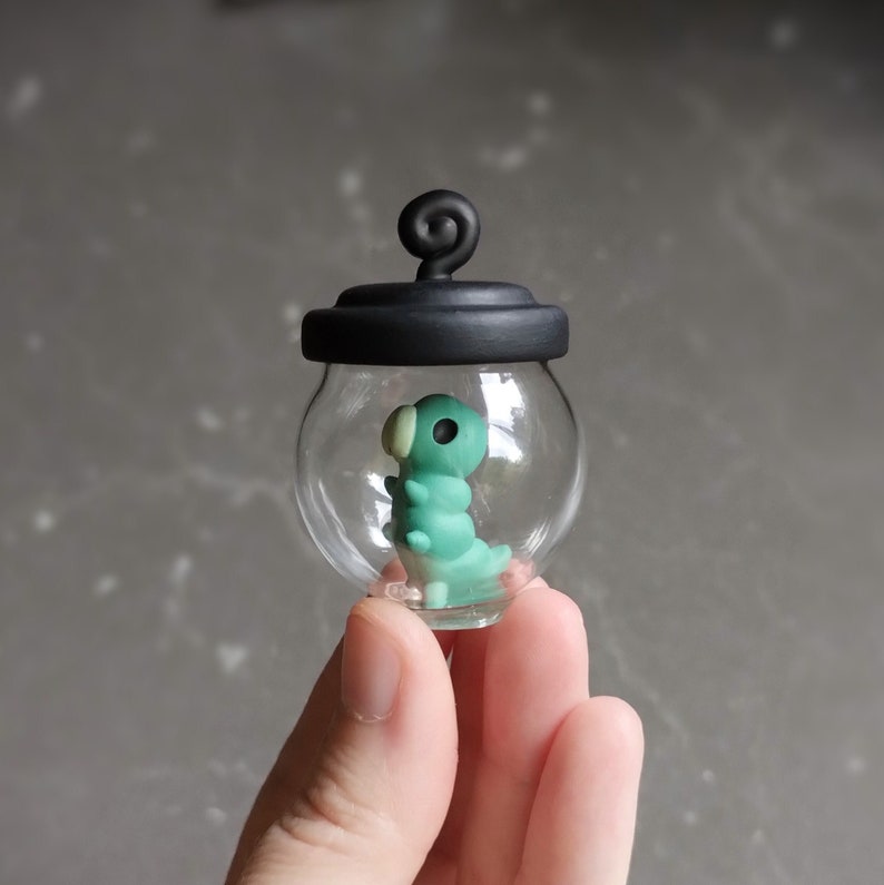Op de afbeelding: Een klein glazen potje met een zwart deksel en een groene en witte rups erin. Het potje wordt in een hand gehouden.