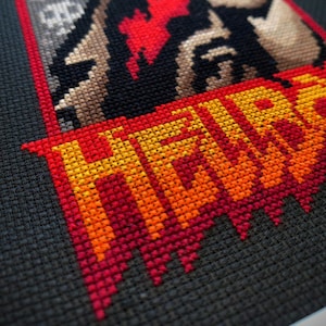 May include: Cross-stitch artwork of a red-skinned character with a cigarette in his mouth, wearing a black leather jacket and a white shirt. The character is standing in front of a red, orange, and yellow cross-stitched word "HELLBOY".