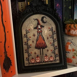May include: A framed cross-stitch featuring a witch with a hat, dress, and ghosts, surrounded by pumpkins and floral accents. The frame is black, and the artwork is displayed on a shelf with other Halloween-themed decor, including a pumpkin and a pillow.