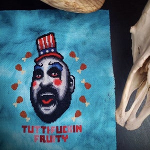 May include: Cross-stitch pattern featuring a portrait of a clown with a red, white, and blue hat, surrounded by a circle of chicken legs. The text "TUTTI FRUTTI" is below the portrait.