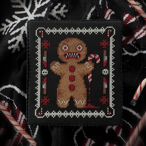 May include: A black and white cross-stitch design of a gingerbread man with a menacing expression holding a candy cane. The gingerbread man is bleeding from a wound on its leg. The design is framed in a black and white border.
