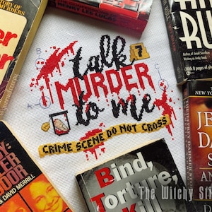 May include: Cross-stitch embroidery with red, black, and yellow thread. The text reads "Talk Murder to Me" with a yellow and black "Crime Scene Do Not Cross" banner below. The embroidery is surrounded by several true crime books.