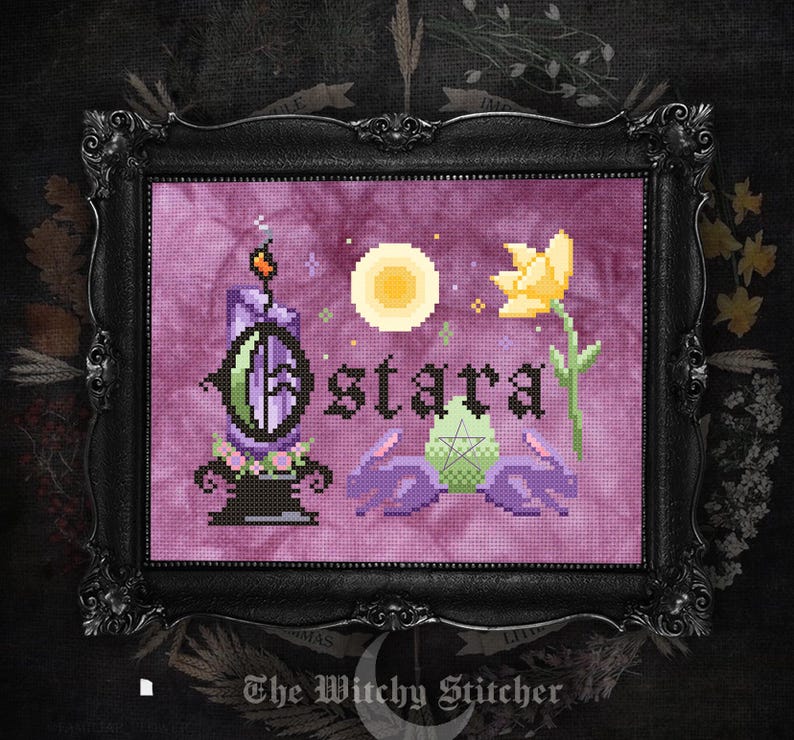 May include: A cross-stitch embroidery in a black ornate frame featuring the text Ostara in black letters. The design includes a purple candle, a yellow flower, a pale green egg with a pentagram, and two purple rabbits against a purple background.