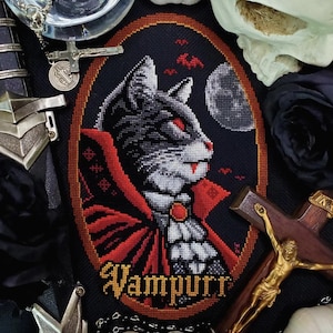 Puede incluir: Patrón de punto de cruz de un gato blanco y negro vestido de vampiro con una capa roja y una joya roja en el cuello. El gato mira hacia la derecha. El fondo es negro con luna llena y murciélagos. El texto "Vampurr" está debajo del gato.