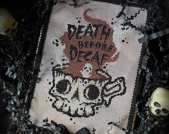 May include: A cross-stitch embroidery on light fabric featuring a skull-shaped coffee mug with steam rising, the text "DEATH BEFORE DECAF," and small ghost figures. The piece is framed with black trim and a small black ribbon bow at the bottom.