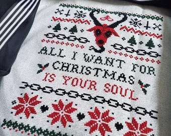 May include: A white fabric cross-stitch piece featuring a red horned creature, green trees, and red snowflakes. The text reads ALL I WANT FOR CHRISTMAS IS YOUR SOUL in black and red lettering. The design includes black chain motifs and holly.