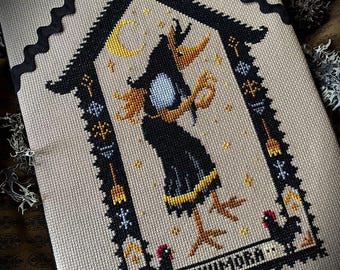 May include: A cross-stitch embroidery on beige fabric featuring a central figure with a bird-like head and black dress, surrounded by a black border with small roosters, stars, and the text KIKIMORA. The piece is accented with black rickrack trim.
