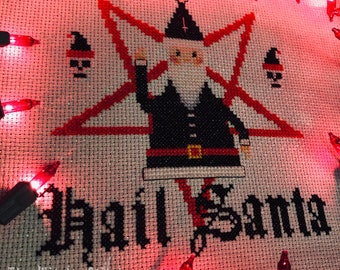 May include: A cross-stitch embroidery on white fabric featuring a central figure of a bearded person in a black suit and hat standing before a large red star. The text "Hail Santa" is stitched in black below, with two small skull icons.