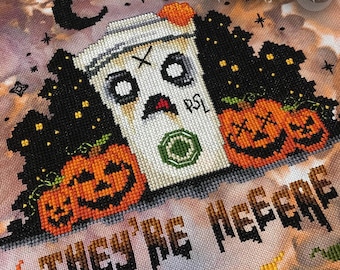Könnte beinhalten: Kreuzstichmuster mit einer weißen Kaffeetasse mit einem Geistergesicht und dem Text "PSL" an der Seite. Vor der Tasse befinden sich drei orangefarbene Kürbisse mit Jack-o'-Lantern-Gesichtern. Der Text "They're Here" befindet sich am unteren Rand des Musters.
