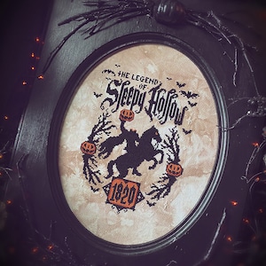 May include: A black framed oval embroidery hoop with a beige background. The embroidery depicts a black silhouette of a rider on horseback with a pumpkin in hand. The text "The Legend of Sleepy Hollow" and "1820" are embroidered in black.