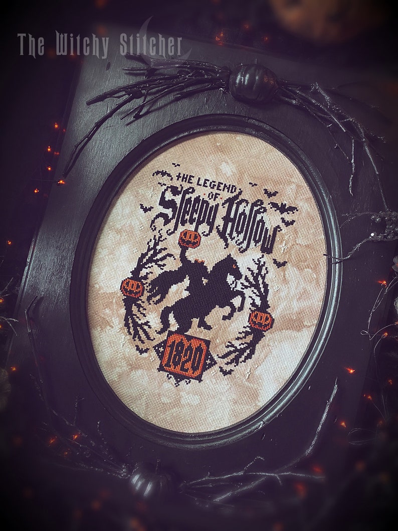 May include: A black framed oval embroidery hoop with a beige background. The embroidery depicts a black silhouette of a rider on horseback with a pumpkin in hand. The text "The Legend of Sleepy Hollow" and "1820" are embroidered in black.