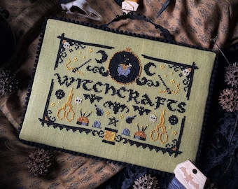 May include: A rectangular cross-stitch sampler on light green fabric with black trim, featuring the text "WITCHCRAFTS" surrounded by embroidered motifs of scissors, buttons, bats, stars, and a cauldron, with a black pom-pom border.