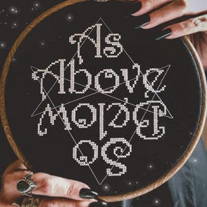 Könnte beinhalten: Ein runder Holzstickrahmen mit schwarzem Stoff und weißem Kreuzstich-Text, der As Above So Below lautet. Ein dünner weißer geometrischer Sternumriss ist hinter dem Schriftzug gestickt. Der Text The Witchy Stitcher ist zu sehen.