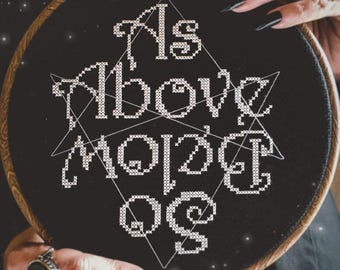 May include: A circular wooden embroidery hoop containing black fabric with white cross-stitched text that reads As Above So Below. A thin white geometric star outline is stitched behind the lettering. The text The Witchy Stitcher is visible.