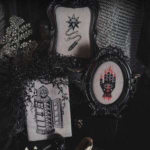 May include: Three framed cross-stitch embroideries titled Relics of the Dark Ages. One shows a flail, one a hand with candles, and one a tower. The frames are ornate black plastic. The text The Witchy Stitcher is visible at the bottom right.