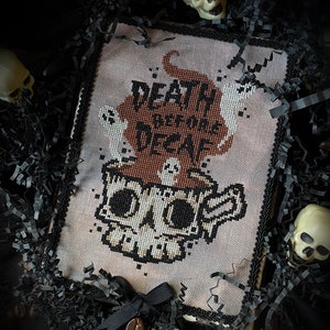 Puede incluir: Arte de punto de cruz con un diseño de taza de café con forma de calavera, con el texto "Death Before Decaf". La obra de arte incluye motivos de fantasmas y calaveras, una cinta negra y un dije de grano de café. La pieza está enmarcada y rodeada de papel negro decorativo y pequeñas calaveras.