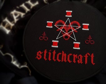 May include: A black embroidery hoop containing a cross-stitch design on black fabric. The design features a red goat head inside a white pentagram, surrounded by red symbols and the red text stitchcraft. The background shows blurred text.