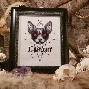 K&ouml;nnte beinhalten: Ein gerahmtes Kreuzstich-Design mit einer schwarzen Katze mit roten Augen, einem Pentagramm und dem Text "Lucifpurr". Der Rahmen ist schwarz und der Hintergrund ist wei&szlig;. Das Bild ist von Tiersch&auml;deln umgeben.