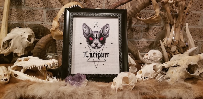 K&ouml;nnte beinhalten: Ein gerahmtes Kreuzstich-Design mit einer schwarzen Katze mit roten Augen, einem Pentagramm und dem Text "Lucifpurr". Der Rahmen ist schwarz und der Hintergrund ist wei&szlig;. Das Bild ist von Tiersch&auml;deln umgeben.