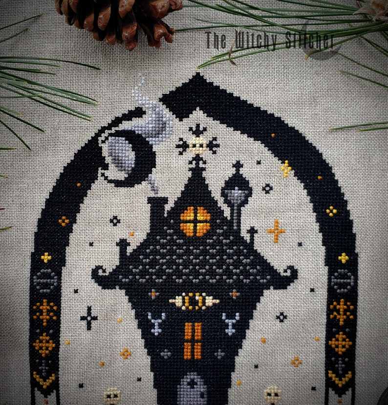May include: Cross stitch pattern featuring a black and gray spooky house with a yellow window, a crescent moon, and stars. The pattern is on a beige linen fabric. The text "The Witchy Stitcher" is in the top right corner.