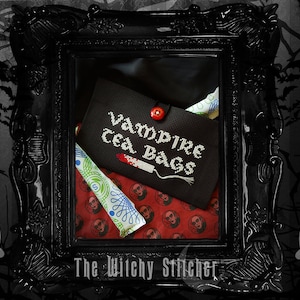 May include: A black fabric pouch with the text "Vampire Tea Bags" embroidered on it. The pouch is open and a white and green tea bag is visible inside. The pouch is in a black ornate frame with a red button.